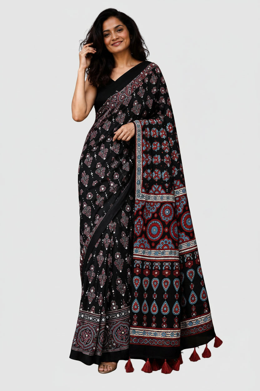 A woman modeling a black saree with intricate red, purple, and white floral and geometric patterns, featuring a solid black blouse and a draped pallu with tassels, set against a plain white background.