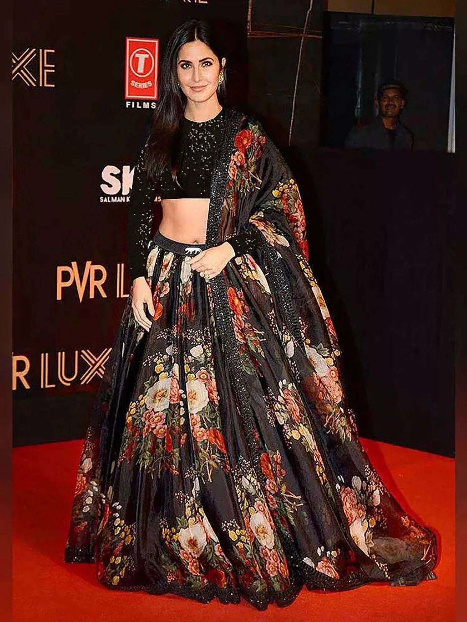 Person wearing a floral and black outfit on a red carpet with logos in the background